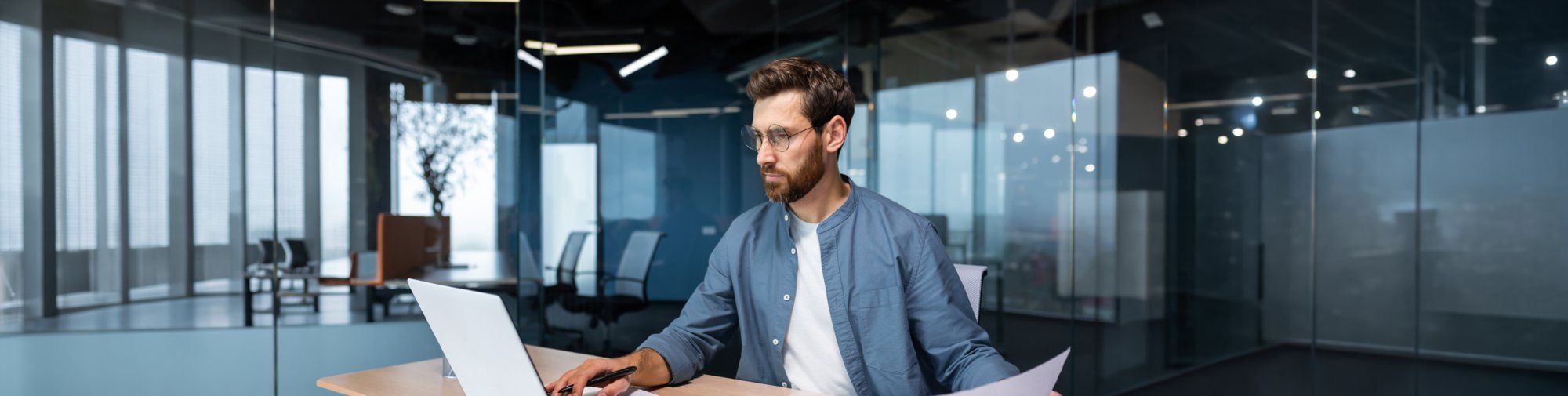 Mann arbeitet in einem modernen Büro an einem Laptop, hält ein Dokument in der Hand und blickt konzentriert auf den Bildschirm.